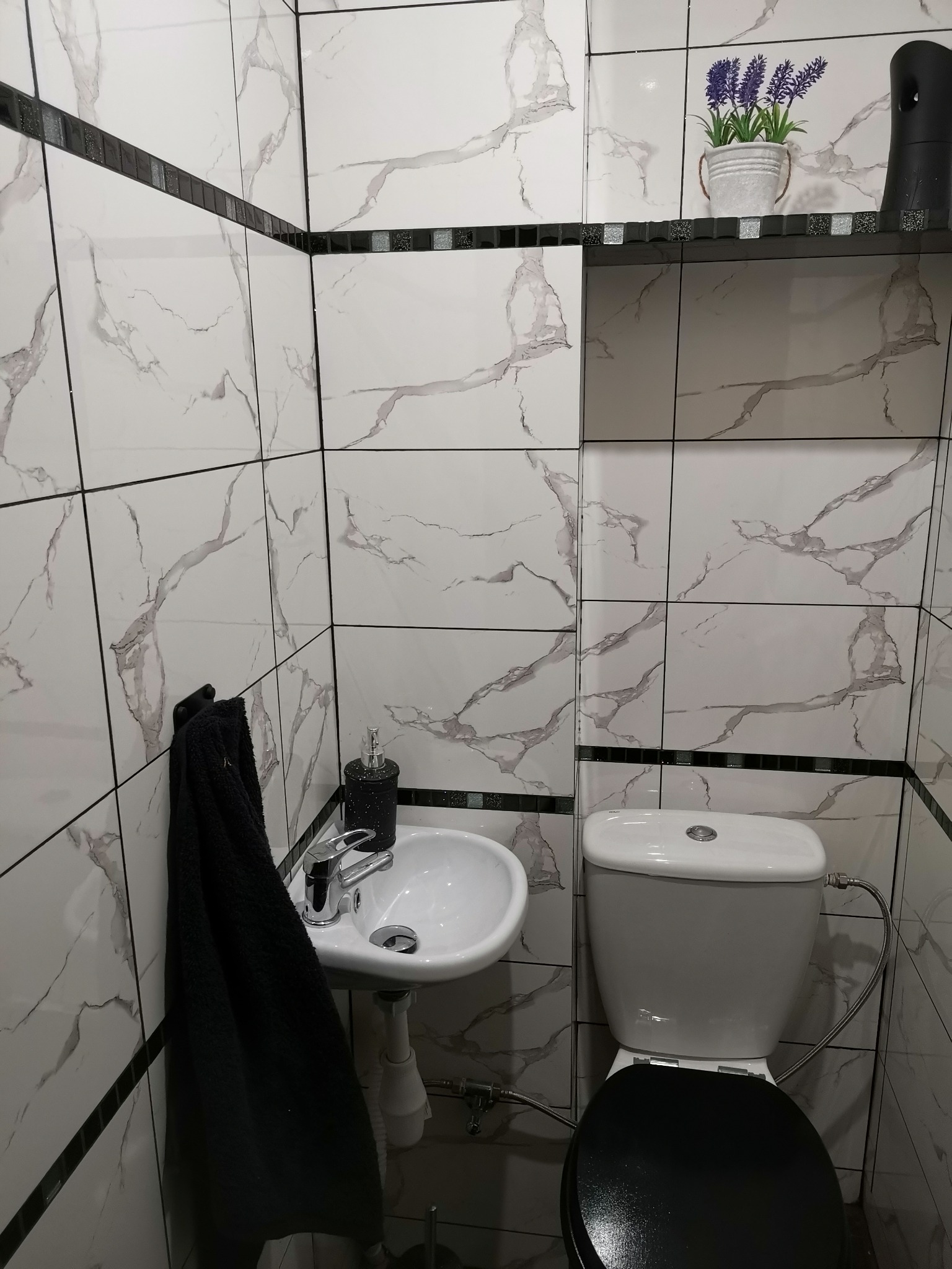 Ukończona łazienka z białymi płytkami imitującymi marmur, czarnym paskiem dekoracyjnym, wiszącą umywalką, toaletą z czarną deską, czarnym ręcznikiem i dekoracją w postaci lawendy w doniczce na półce.