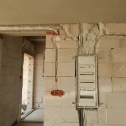 Rozdzielnica elektryczna z licznymi kablami w ścianie z pustaków, widoczne przewody prowadzone w korytach i peszlach, instalacja w trakcie montażu w surowym stanie.