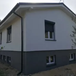 Dwukondygnacyjny dom z jasnoszarą elewacją i ciemnoszarą podmurówką, widoczne okna z roletami zewnętrznymi i system rynnowy.