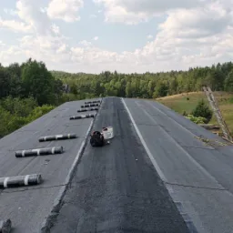 Dach pokryty papą termozgrzewalną w trakcie prac dekarskich, widoczne rolki papy, wiadro z klejem, drabina oparta o dach i malowniczy krajobraz z lasem w tle.