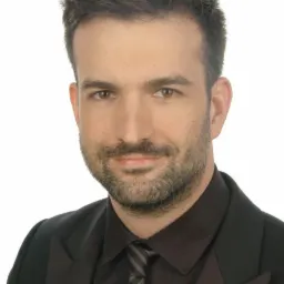 Portret brodatego mężczyzny w ciemnej koszuli i marynarce z szaro-czarnym krawatem na białym tle.