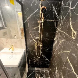 Nowoczesna kabina prysznicowa z czarnymi marmurowymi płytkami z białymi żyłami, złota armatura, deszczownica i bateria, umywalka z białą ceramiką i złotym kranem widoczne za szklaną ścianką.