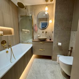 Refit Solutions Paweł Suchocki - Beżowa łazienka z wanną, umywalką na szafce i toaletą. Złote detale armatury i oświetlenia. Płytki w odcieniach beżu i szarości. Nowoczesny design z podświetleniem LED.