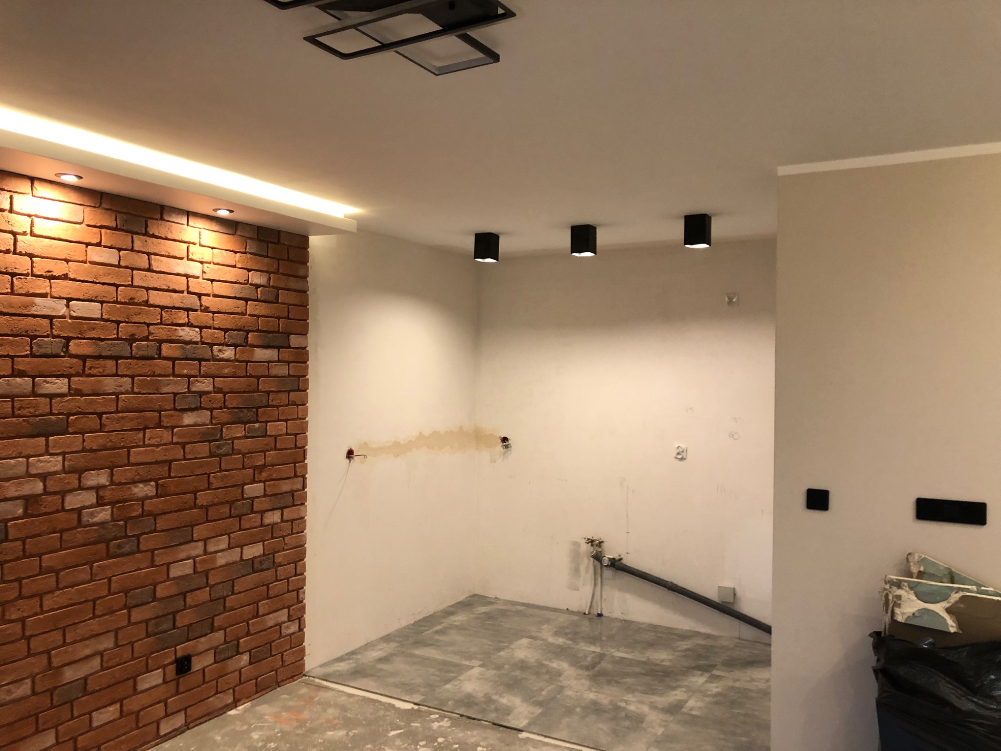 Wnętrze w trakcie prac wykończeniowych: ściana z cegły klinkierowej z oświetleniem LED, surowa podłoga z płytek, instalacja wodna i elektryczna w stanie surowym, widoczne punkty świetlne na suficie.