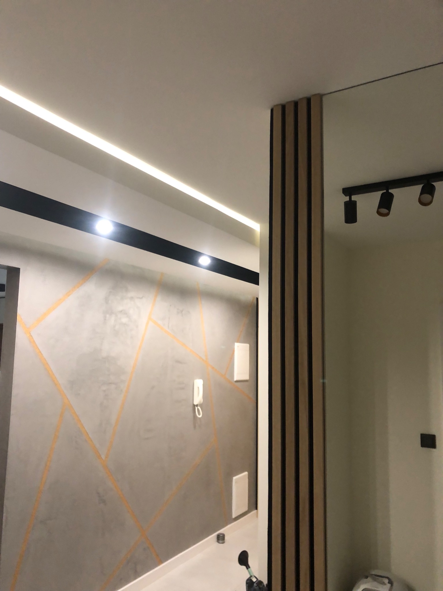Nowoczesny korytarz z akcentem geometrycznym na szarej ścianie, listwami dekoracyjnymi z drewna, oświetleniem LED i reflektorami punktowymi.