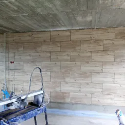Ściana wyłożona płytkami imitującymi drewno, widoczna przecinarka do płytek, surowy betonowy sufit, instalacja wodna w tle.