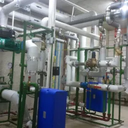 Rozbudowana instalacja z rur, zaworów, pomp i zbiorników w kotłowni, z przewagą kolorów białego, szarego i zielonego.