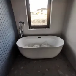 Nowa, wolnostojąca wanna w minimalistycznej łazience z oknem, jeszcze zabezpieczona folią ochronną wewnątrz, z widoczną chromowaną baterią podłogową i szarą, cegiełkową okładziną ścienną ułożoną...