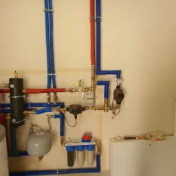 Rozbudowana instalacja centralnego ogrzewania z rurami w izolacji, zbiornikiem wyrównawczym, filtrami wody i pompami obiegowymi zamontowana na ścianie w pomieszczeniu technicznym.