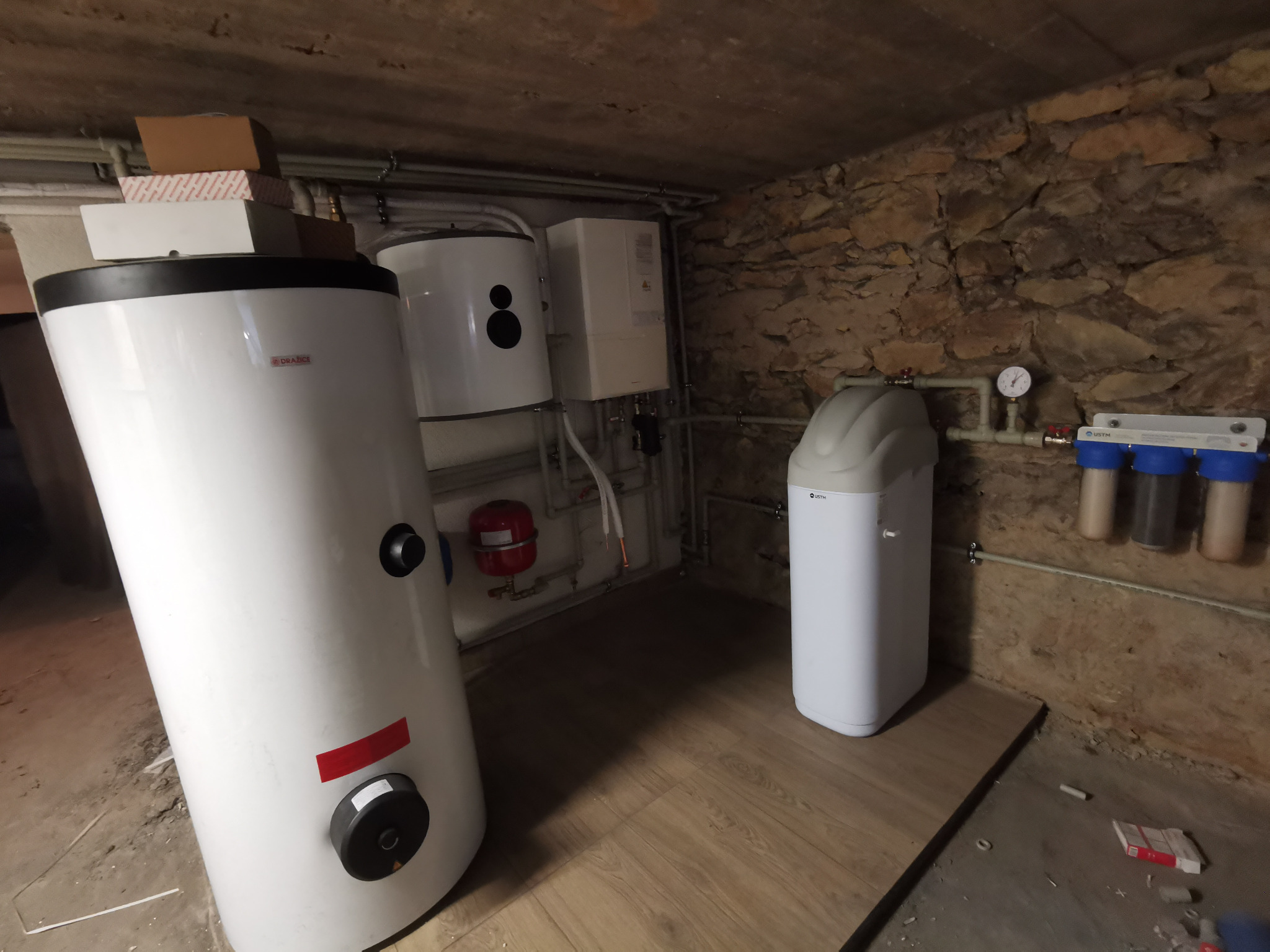 Instalacja kotłowni w piwnicy z kamienną ścianą: biały bojler marki Drazice, mniejszy bojler, ekspansomat, zmiękczacz wody USTM i filtry mechaniczne.