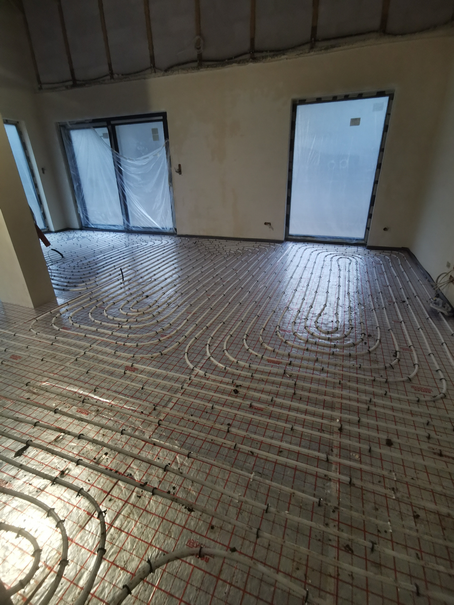 Rozłożona instalacja ogrzewania podłogowego w nowym budynku, rury ułożone na siatce z folią aluminiową, widoczne okna zabezpieczone folią.