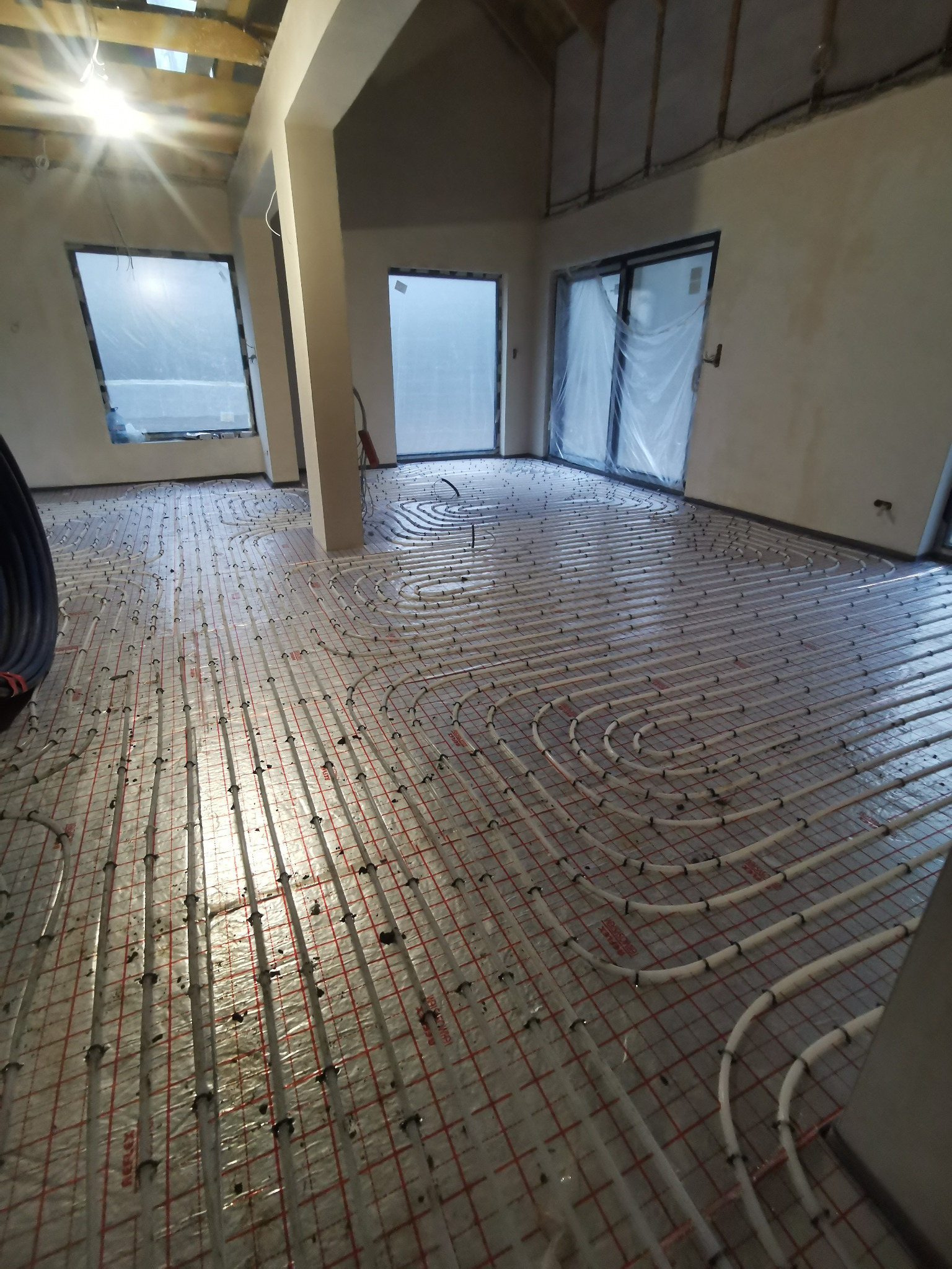 Rozłożone rury ogrzewania podłogowego na siatce w surowym wnętrzu domu jednorodzinnego, widoczne okna i surowy strop.