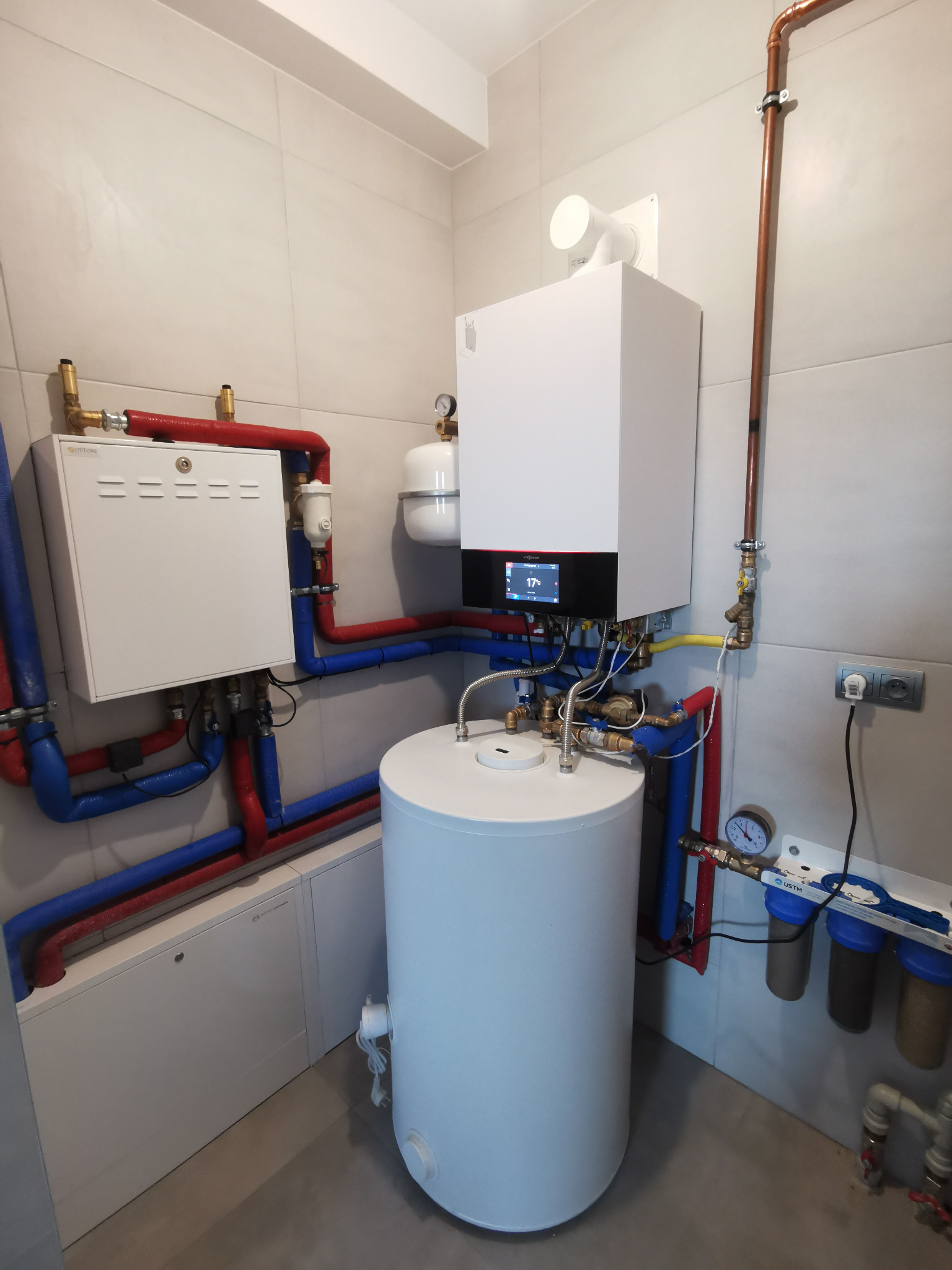 Nowoczesna kotłownia z białym piecem gazowym Viessmann, zasobnikiem ciepłej wody i rozbudowaną instalacją hydrauliczną z rurami w izolacji czerwono-niebieskiej, całość w jasnym pomieszczeniu...