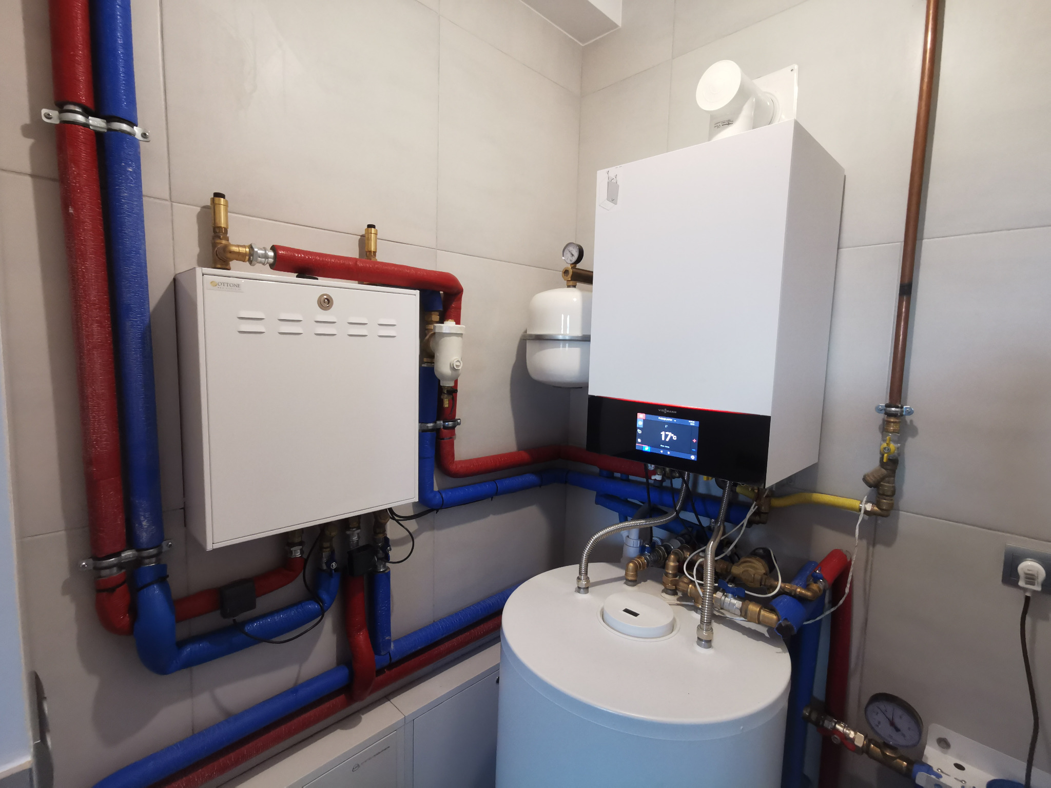 Nowoczesna kotłownia z piecem gazowym, zasobnikiem ciepłej wody i rozdzielnicą, rury w kolorach czerwonym i niebieskim, manometry, instalacja na tle jasnych płytek.