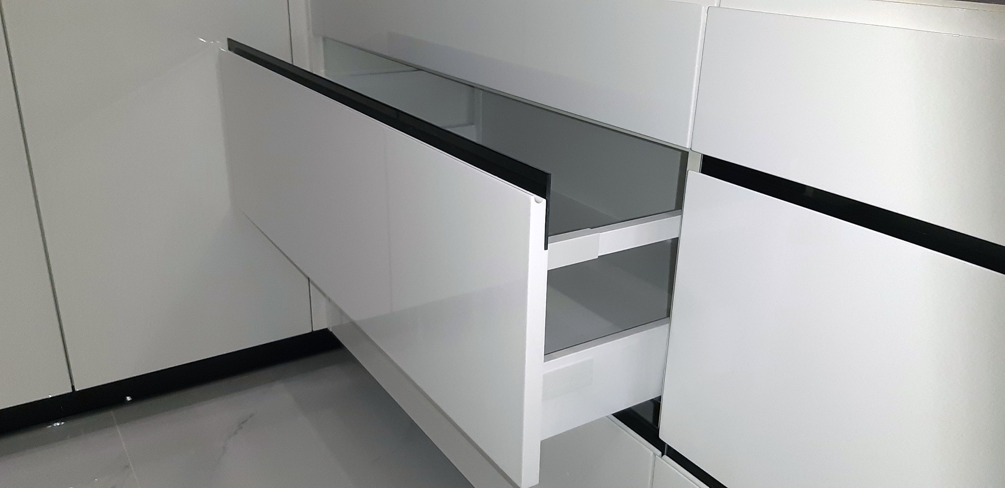 Nowoczesna, biała szafka kuchenna z wysuwanymi szufladami, czarne akcenty na krawędziach, widoczne dwie półki w szufladzie.