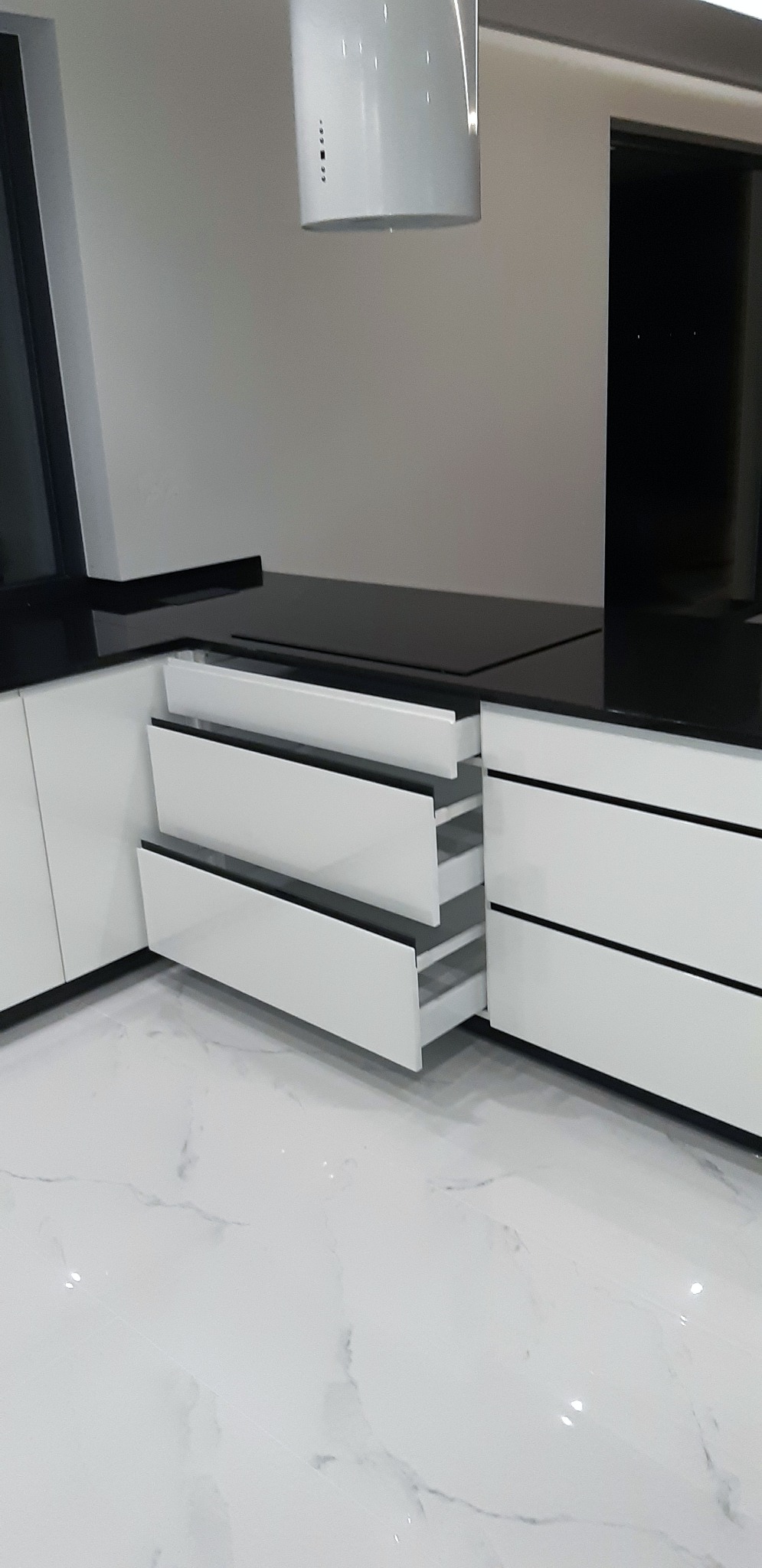 Nowoczesna kuchnia z białymi frontami szafek i szuflad z czarnymi akcentami, otwarte szuflady ukazują minimalistyczne wnętrze, widoczny fragment marmurowej podłogi i okapu.