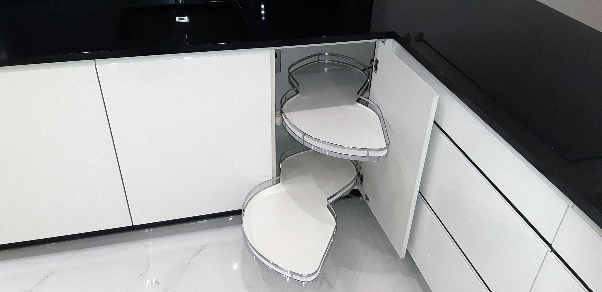 Wysuwana, dwupoziomowa półka narożna w szafce kuchennej z białymi frontami i czarnym blatem, widoczna na tle marmurowej podłogi.