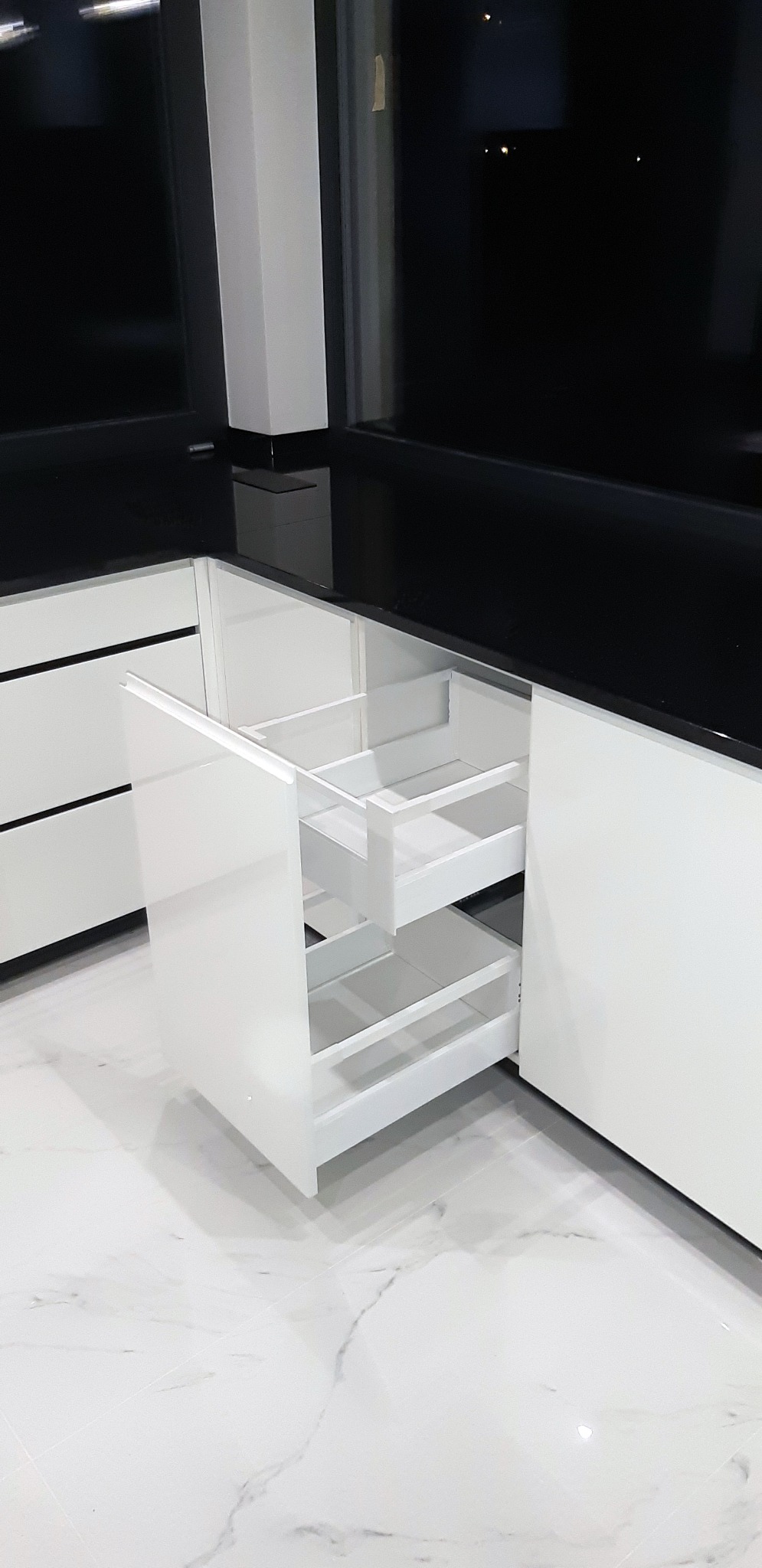 Wysuwana, dwupoziomowa szuflada cargo w białej, nowoczesnej zabudowie kuchennej z czarnym blatem, widok na marmurową podłogę.