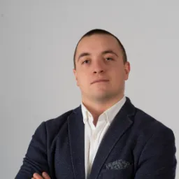 Portret mężczyzny w granatowej marynarce z poszetką w kieszonce, skrzyżowane ręce na piersiach, biała koszula z rozpiętym guzikiem pod szyją na jasnym tle.