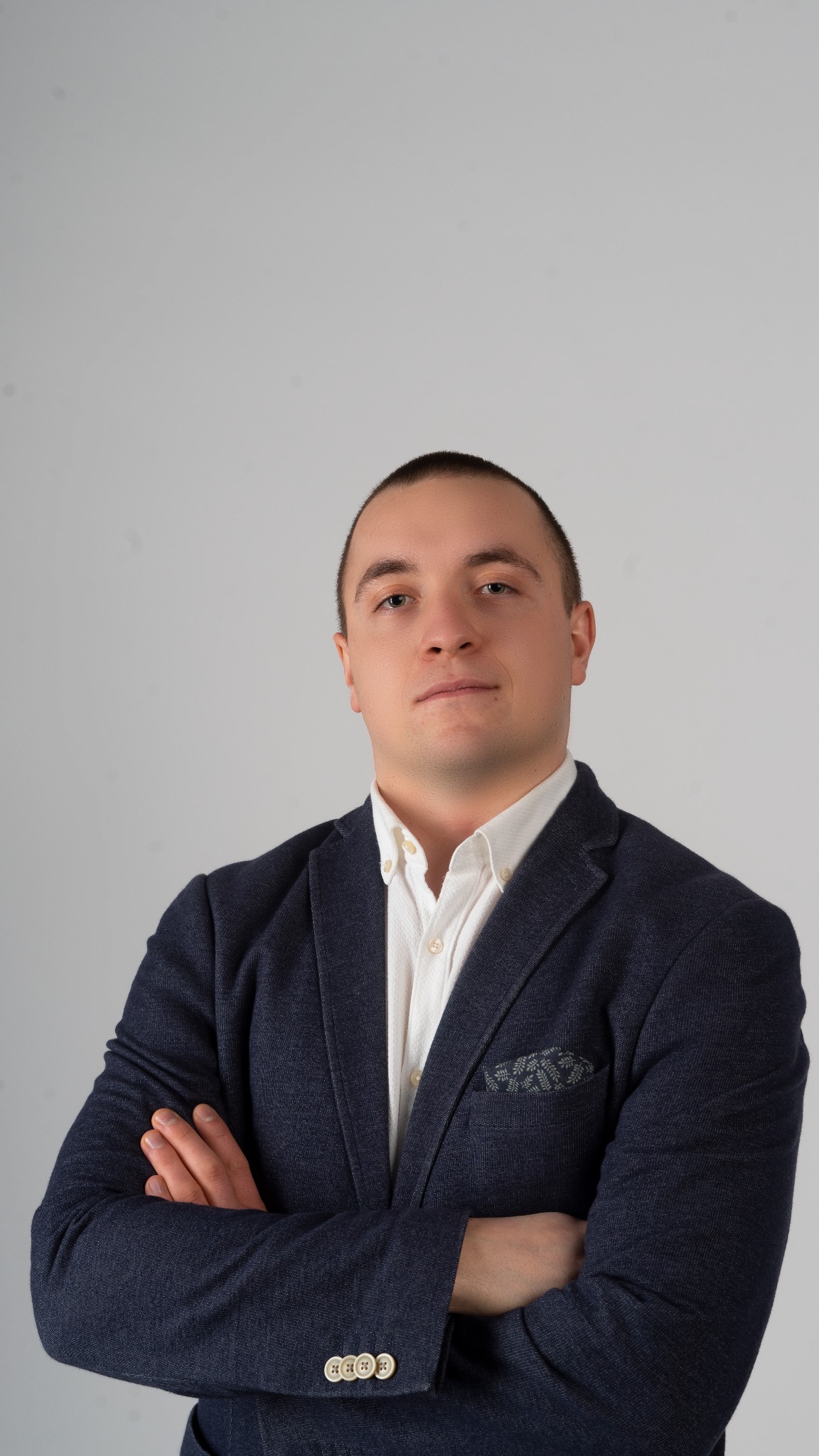 Portret mężczyzny w granatowej marynarce z poszetką w kieszonce, skrzyżowane ręce na piersiach, biała koszula z rozpiętym guzikiem pod szyją na jasnym tle.