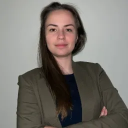 Portret kobiety w oliwkowym żakiecie i granatowej koszulce, skrzyżowane ramiona, długie brązowe włosy, neutralne tło.