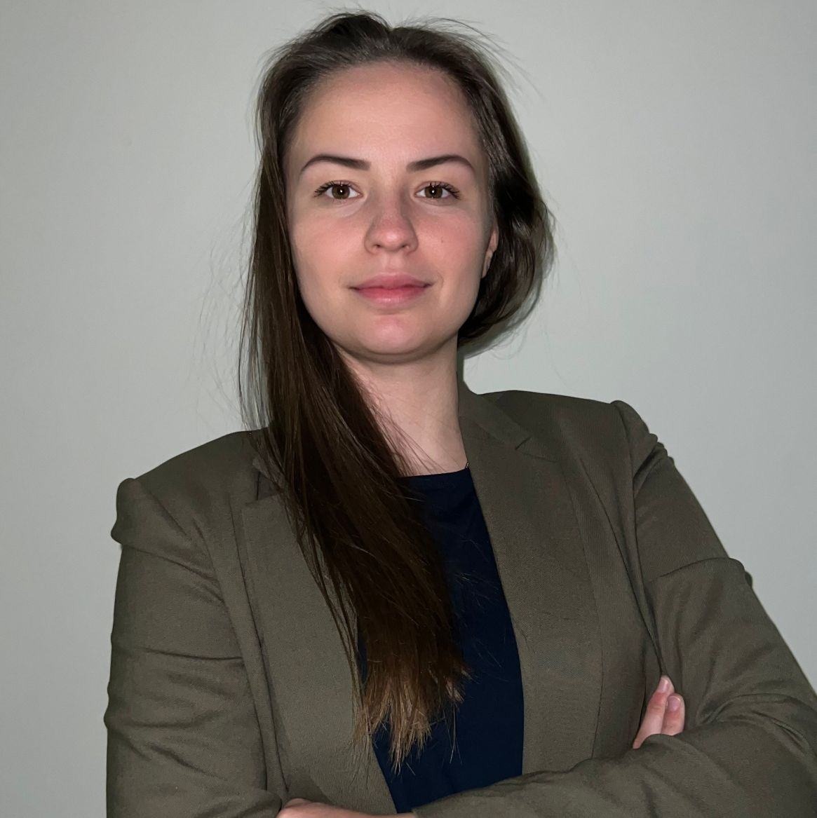 Portret kobiety w oliwkowym żakiecie i granatowej koszulce, skrzyżowane ramiona, długie brązowe włosy, neutralne tło.