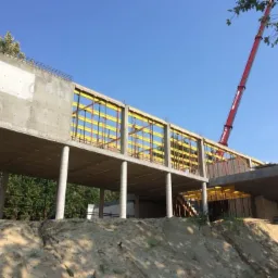 Surowa konstrukcja domu z betonu, widoczne żółte szalunki i czerwony dźwig na tle błękitnego nieba.