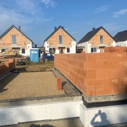 Usługi ogólnobudowlane Arkadiusz Kuczyński - Fundamenty z bloczków betonowych i izolacji, przygotowane pod wznoszenie ścian z czerwonej cegły ceramicznej; w tle budynki w stanie surowym otwarte i przenośna toaleta.