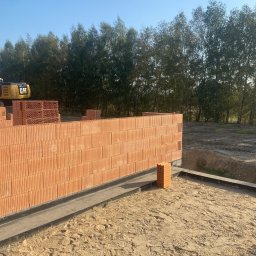 Usługi ogólnobudowlane Arkadiusz Kuczyński - Wznoszenie ściany z czerwonych, porowatych bloczków ceramicznych na fundamencie z hydroizolacją, w tle widoczna koparka CAT i drzewa.