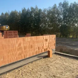 Wznoszenie ściany z czerwonych, porowatych bloczków ceramicznych na fundamencie z hydroizolacją, w tle widoczna koparka CAT i drzewa.