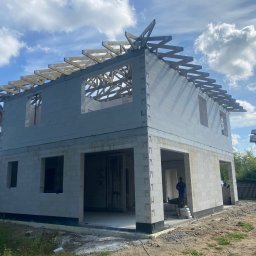 Usługi ogólnobudowlane Arkadiusz Kuczyński - Dwukondygnacyjny budynek w stanie surowym otwartym, wzniesiony z białych bloczków, z widoczną konstrukcją więźby dachowej, na tle błękitnego nieba z chmurami.