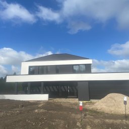 Usługi ogólnobudowlane Arkadiusz Kuczyński - Nowoczesny dom z płaskim dachem w trakcie budowy, z widoczną elewacją w kolorze białym i czarnym, na pierwszym planie sterta piasku i oznaczenia geodezyjne.