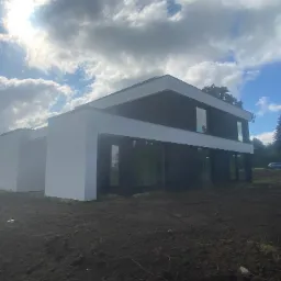 Nowoczesny dom z płaskim dachem, wykończony w bieli i czerni, na tle pochmurnego nieba i świeżo przekopanej ziemi.