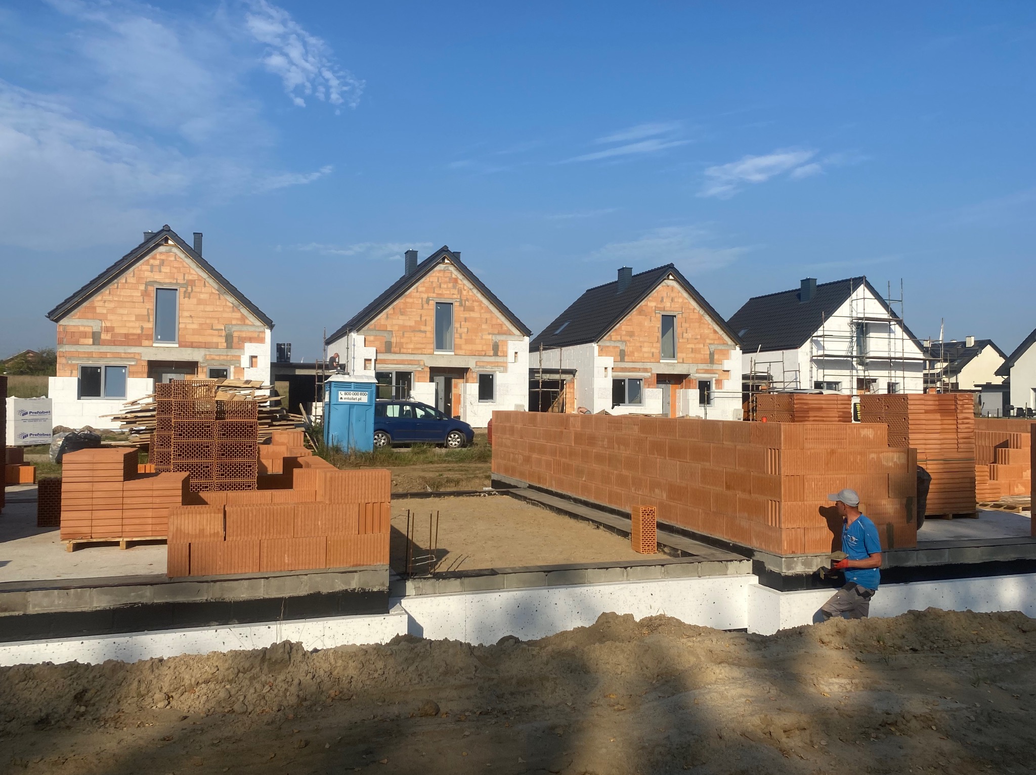 Budowa szeregu domów jednorodzinnych z czerwonej cegły, widoczne fundamenty, materiały budowlane i pracownik w niebieskiej koszulce.