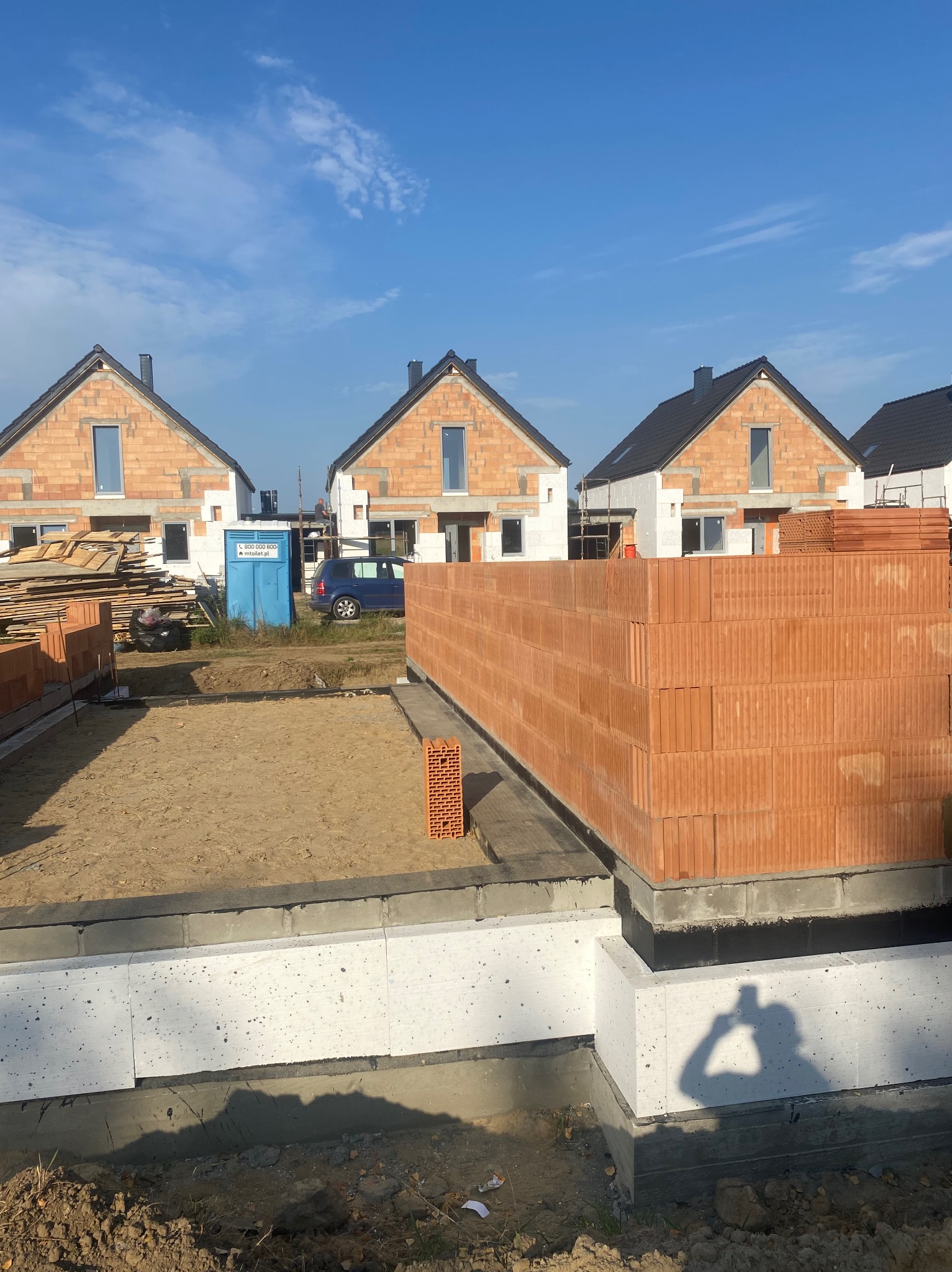 Fundamenty z bloczków betonowych i izolacji, przygotowane pod wznoszenie ścian z czerwonej cegły ceramicznej; w tle budynki w stanie surowym otwarte i przenośna toaleta.