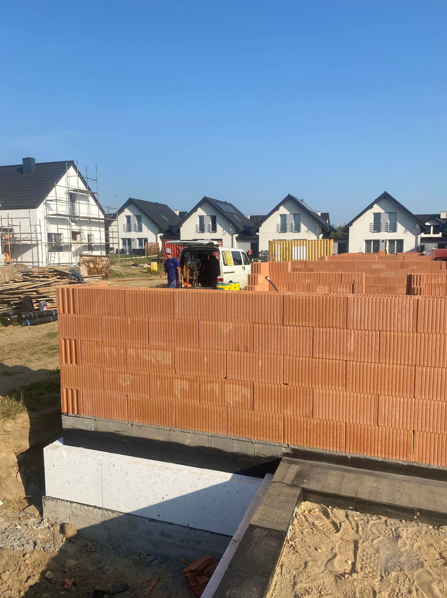 Wznoszenie ściany z czerwonej ceramiki poryzowanej na fundamencie z izolacją, w tle budowa osiedla domów jednorodzinnych i samochód dostawczy.