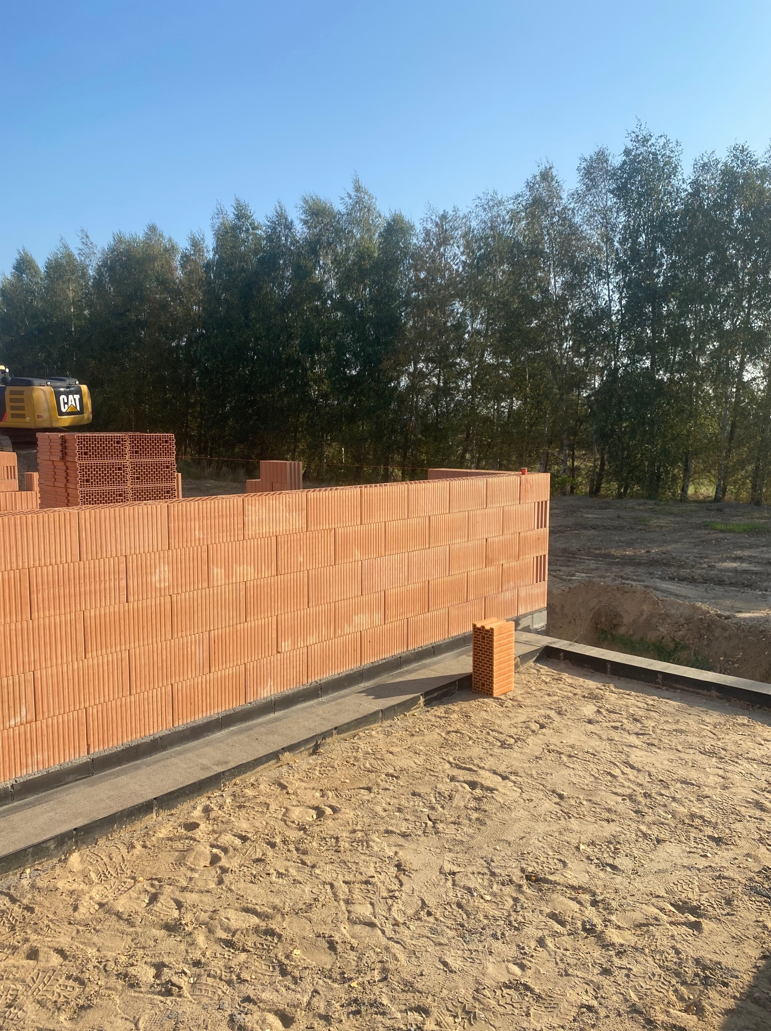 Wznoszenie ściany z czerwonych, porowatych bloczków ceramicznych na fundamencie z hydroizolacją, w tle widoczna koparka CAT i drzewa.