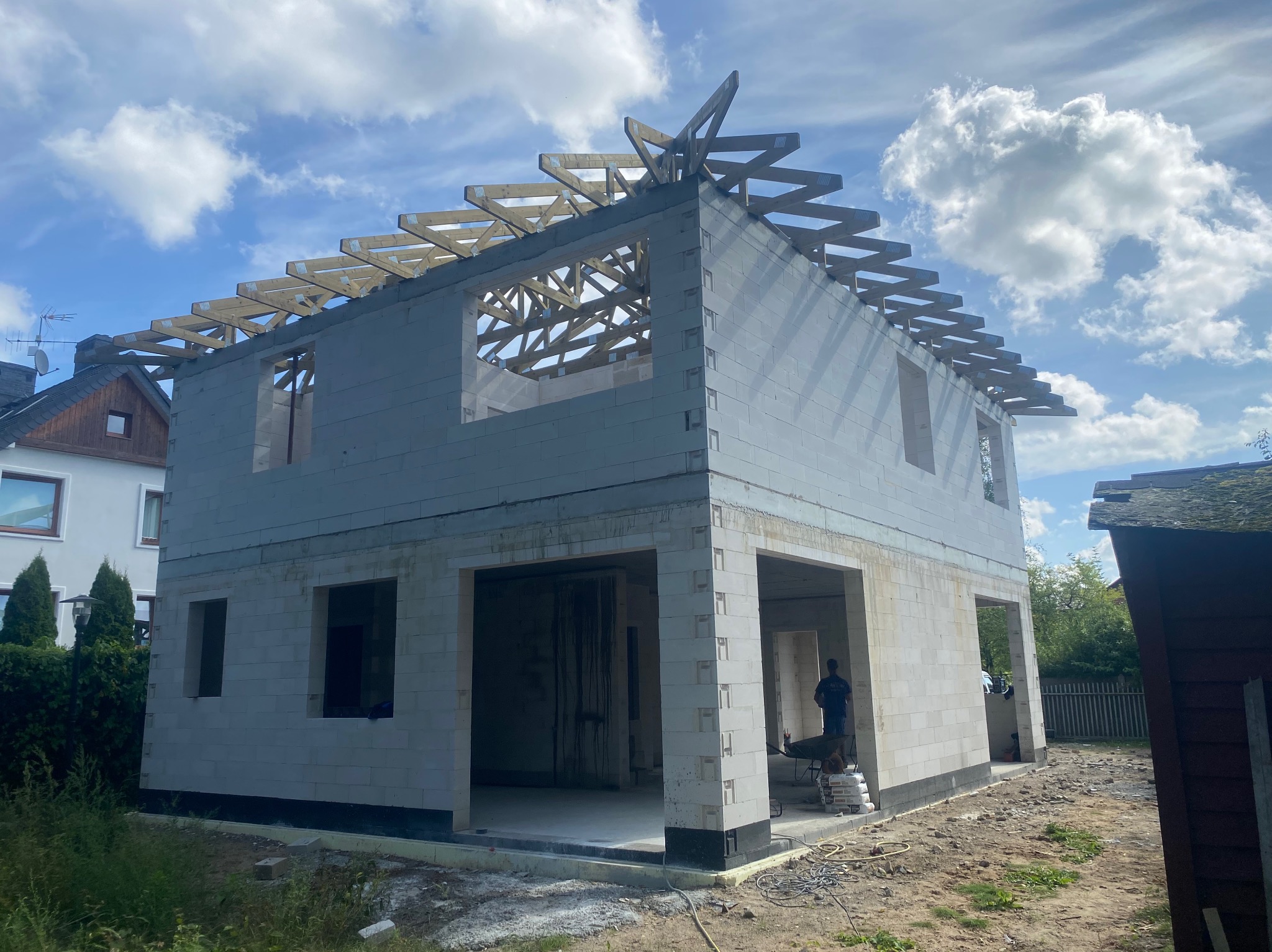 Dwukondygnacyjny budynek w stanie surowym otwartym, wzniesiony z białych bloczków, z widoczną konstrukcją więźby dachowej, na tle błękitnego nieba z chmurami.