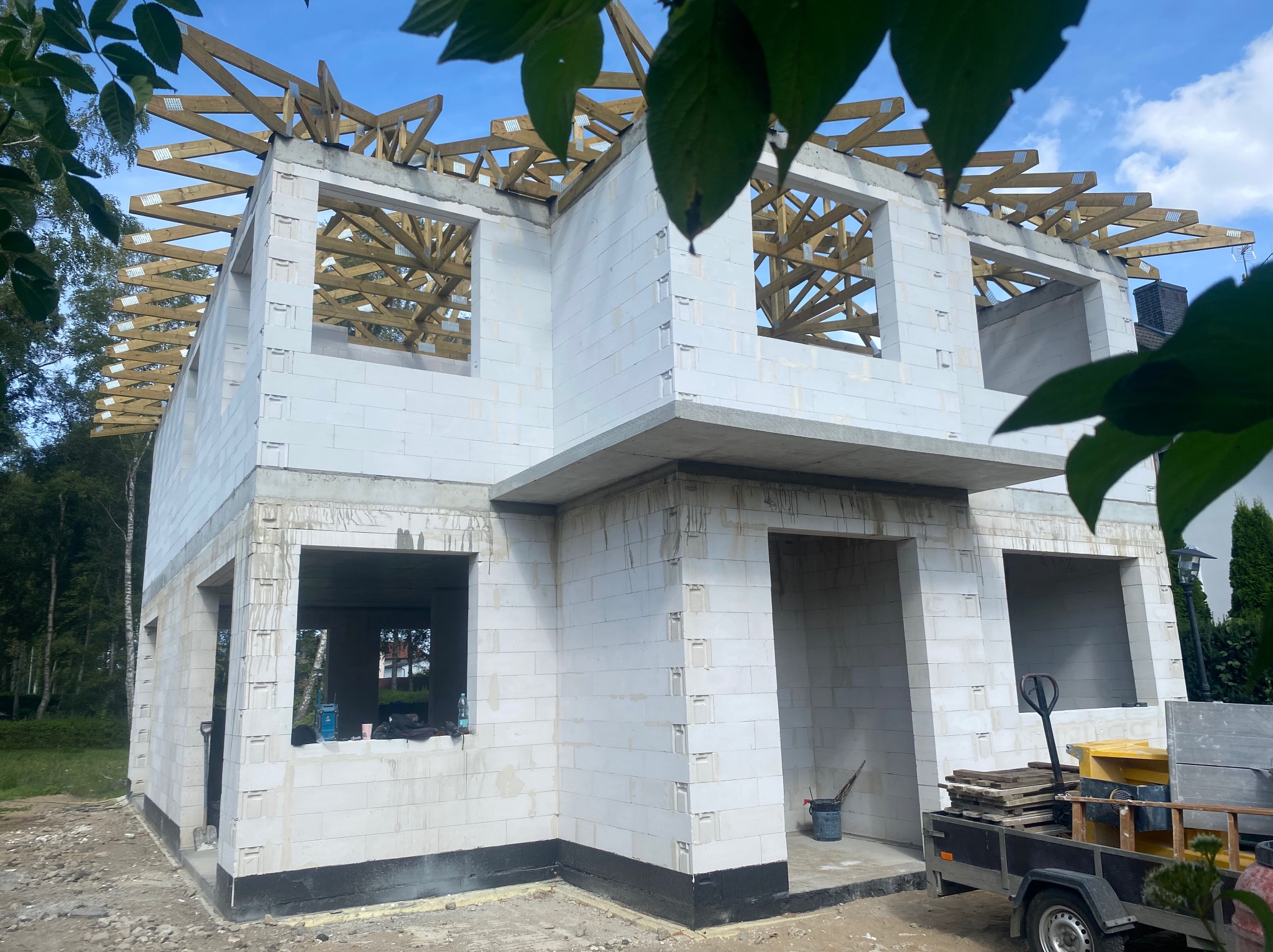 Dwukondygnacyjny dom murowany w stanie surowym otwartym z drewnianą konstrukcją dachu, widoczny z poziomu gruntu, z przyczepką i narzędziami budowlanymi w tle.