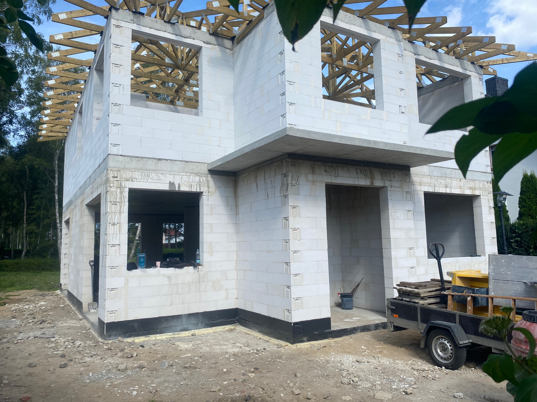 Surowy stan dwukondygnacyjnego domu z białych bloczków, widoczna konstrukcja drewniana dachu, przyczepka z materiałami budowlanymi i narzędziami na placu budowy.