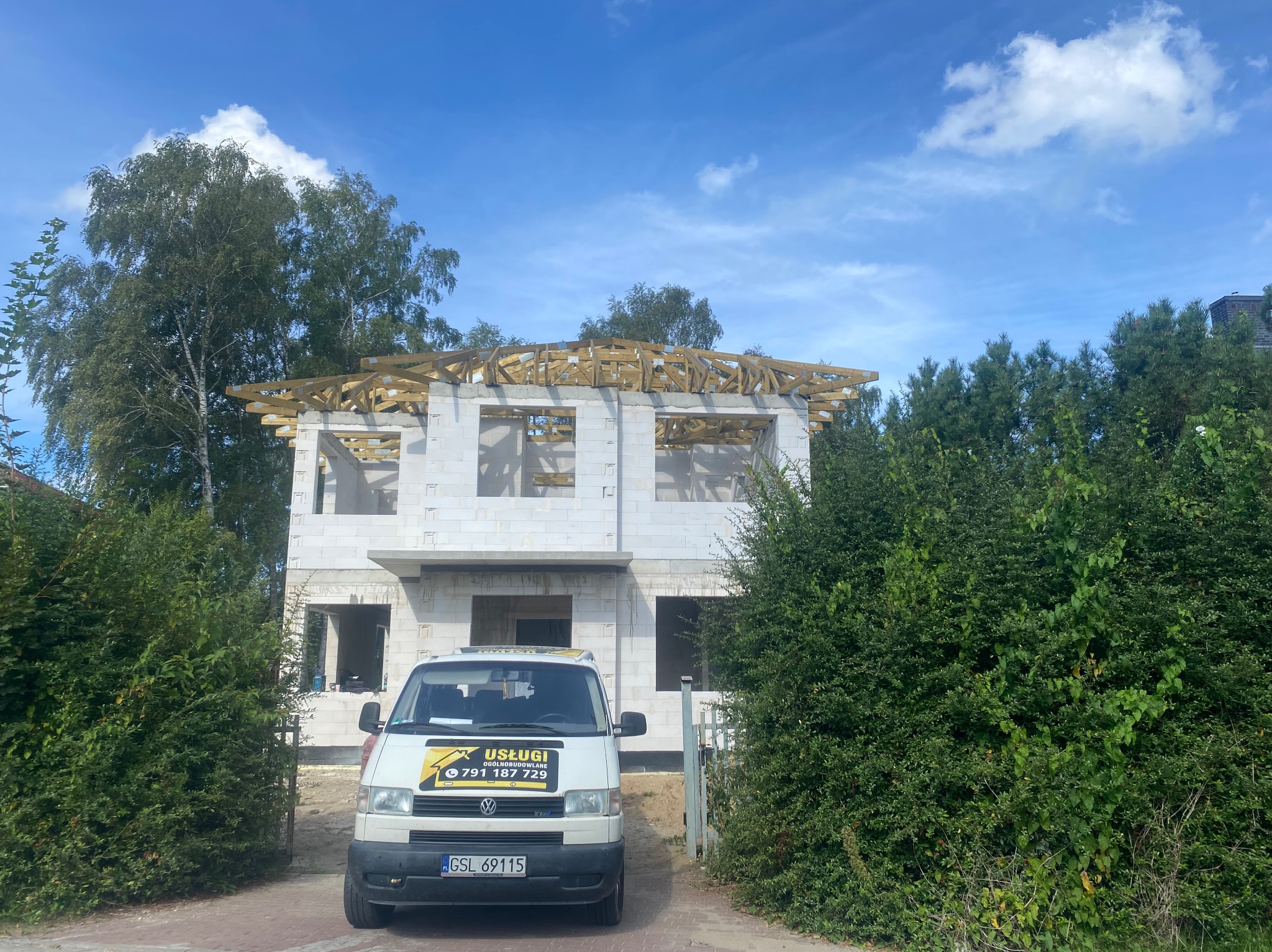 Niedokończony dwukondygnacyjny dom z białego bloczka z drewnianą konstrukcją dachu, na pierwszym planie biały bus z naklejką firmy budowlanej na tle zieleni i błękitnego nieba.