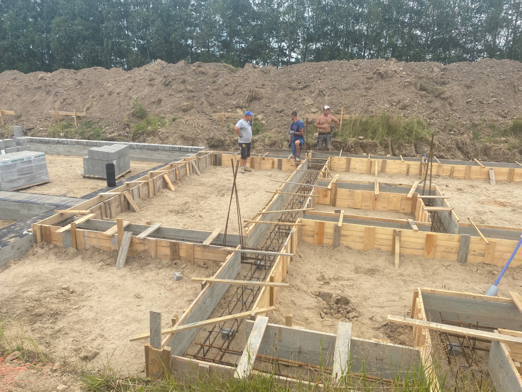 Fundamenty domu w trakcie budowy: drewniane szalunki zbrojone stalą, widoczni trzej robotnicy na tle ziemnego nasypu i zieleni.