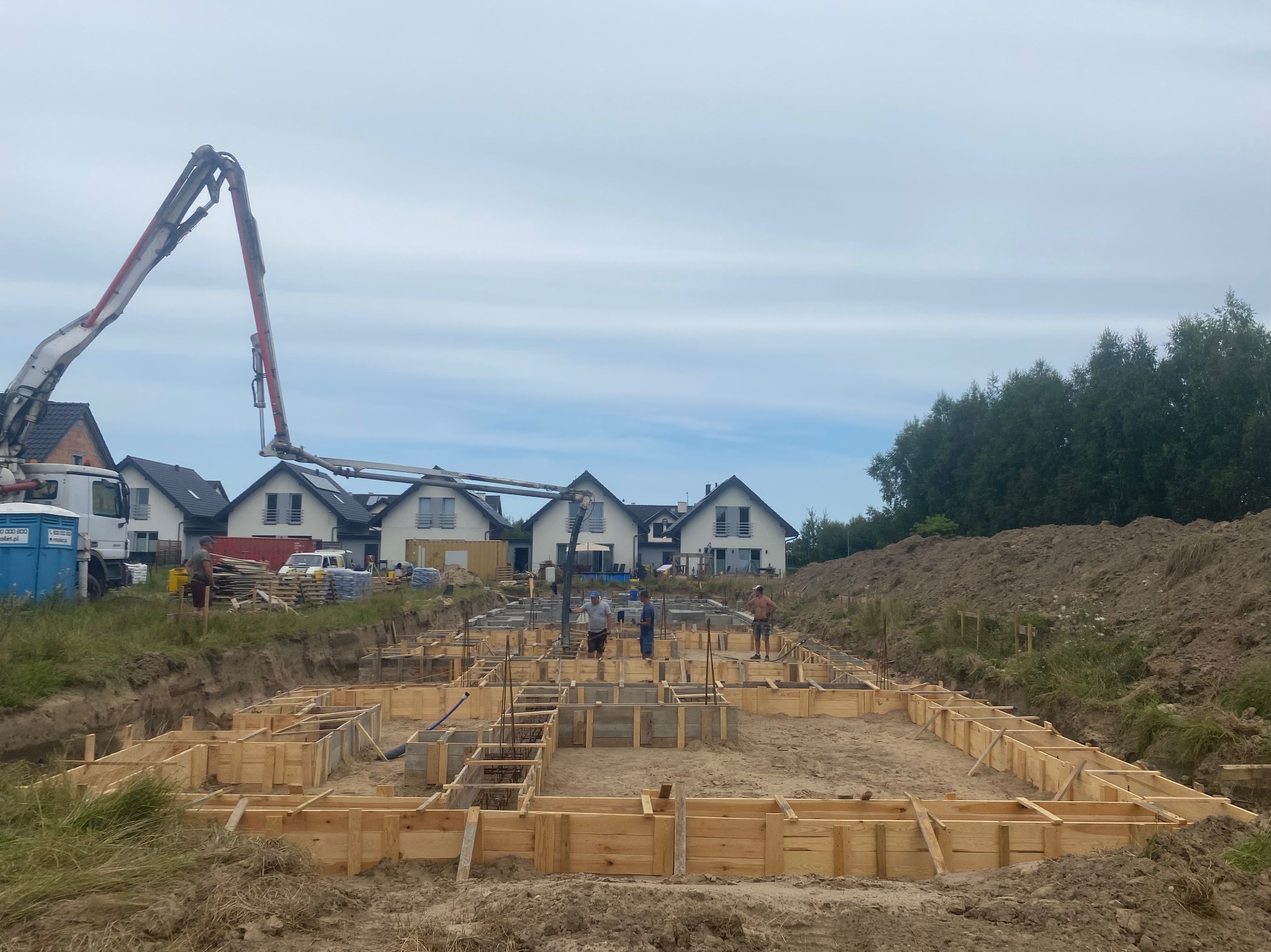 Budowa fundamentów domu jednorodzinnego z użyciem szalunków drewnianych, widoczny betonowóz z pompą i pracownicy na placu budowy.