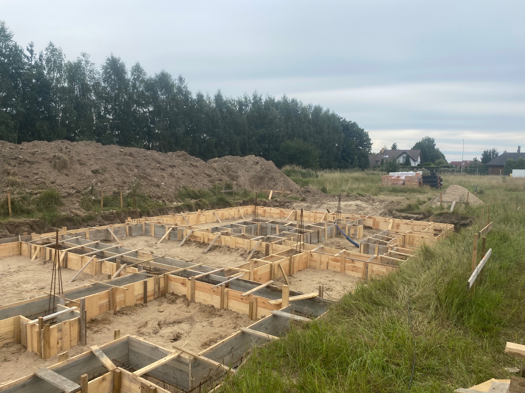 Przygotowanie fundamentów pod budowę domu: drewniany szalunek zbrojony stalą, zalewany betonem, na tle ziemi, trawy i drzew.