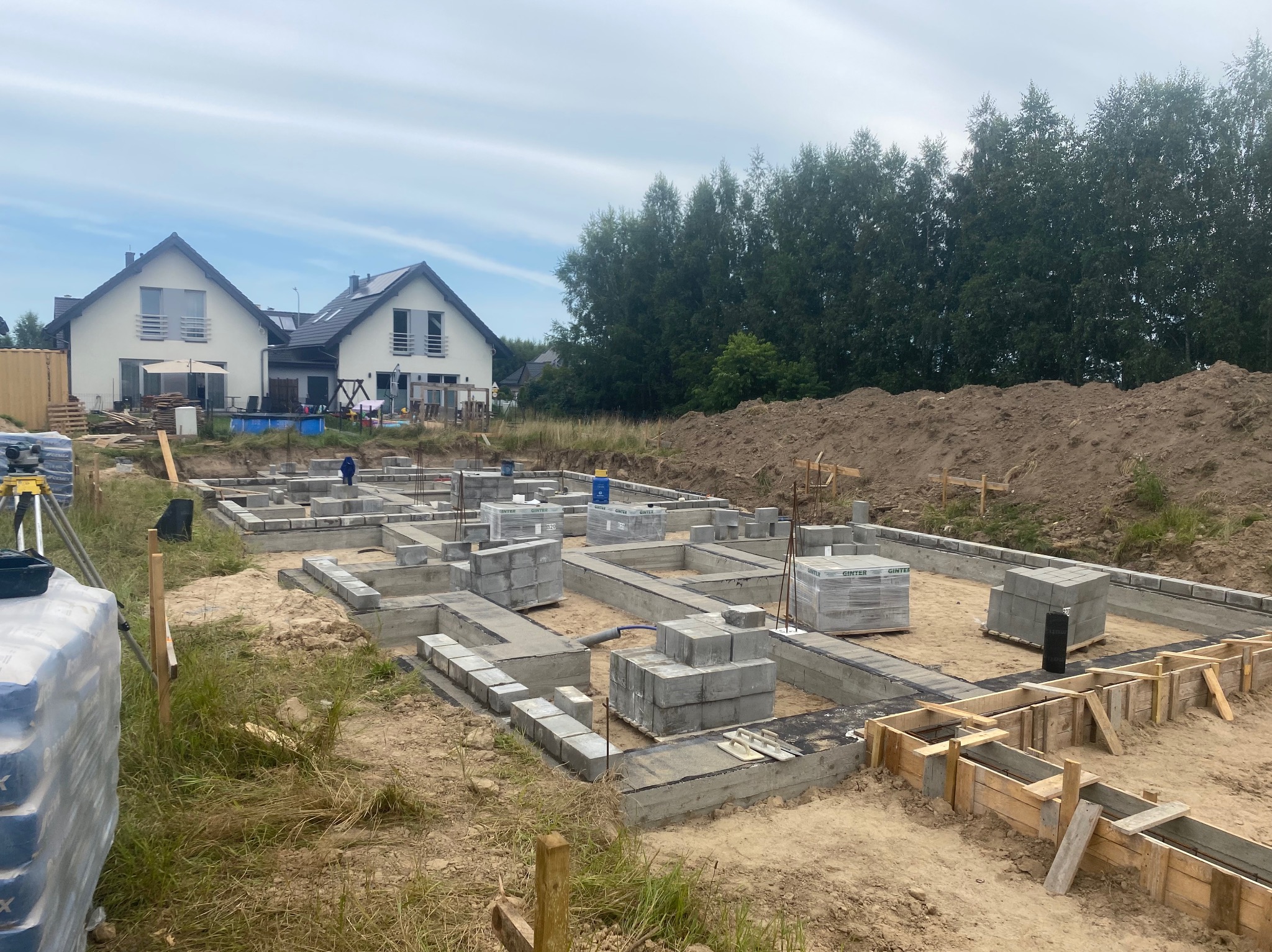 Fundamenty pod budowę domu jednorodzinnego z widocznymi bloczkami betonowymi i szalunkiem, w tle dwa gotowe domy i drzewa.