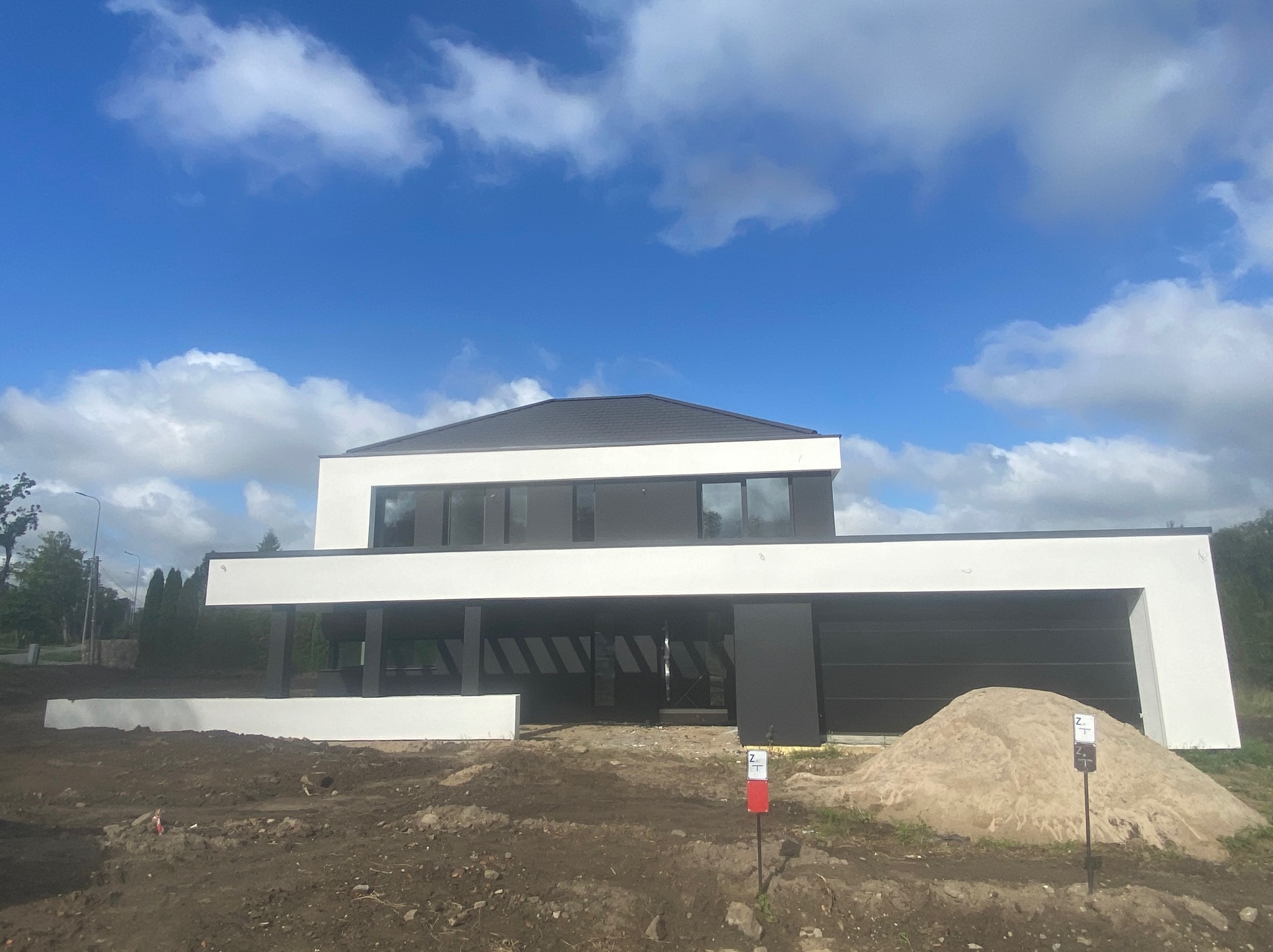 Nowoczesny dom z płaskim dachem w trakcie budowy, z widoczną elewacją w kolorze białym i czarnym, na pierwszym planie sterta piasku i oznaczenia geodezyjne.