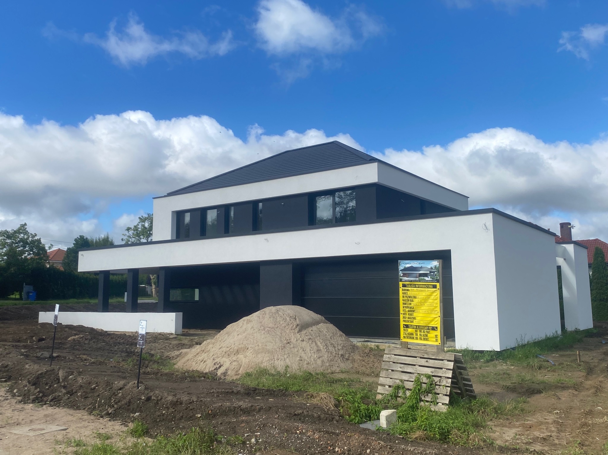 Nowoczesny, dwukondygnacyjny dom murowany w Słupsku, z płaskim dachem i białymi ścianami, w trakcie budowy; widoczna sterta piasku i tablica informacyjna.