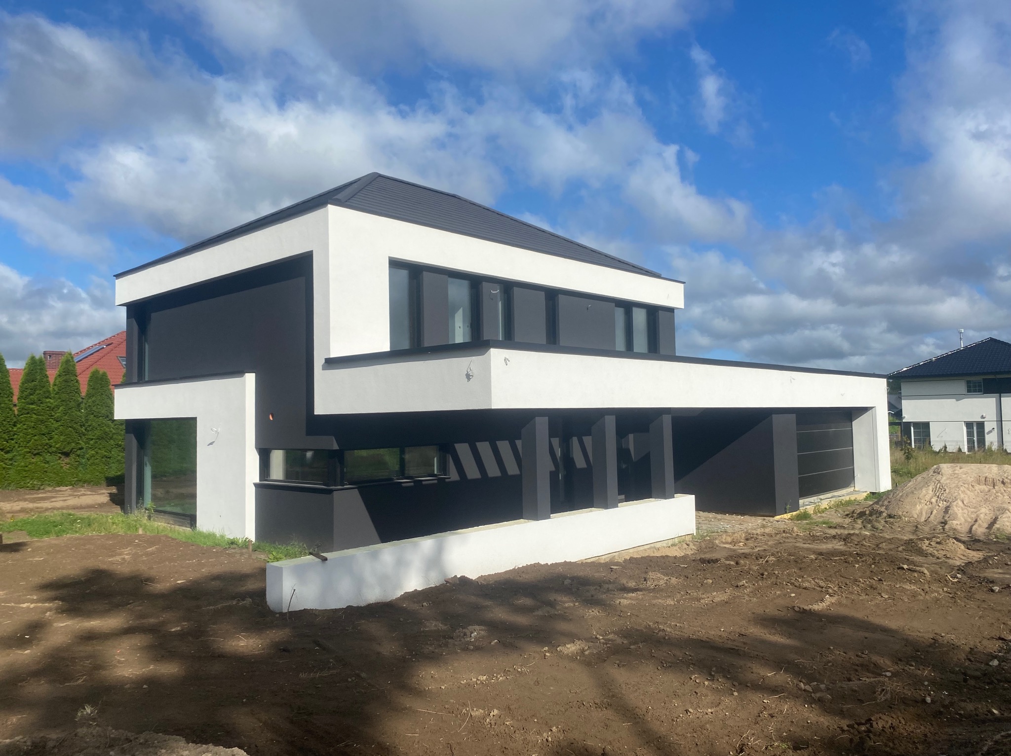 Nowoczesny, dwukondygnacyjny dom murowany z płaskim dachem, wykończony w bieli i czerni, z widocznym fragmentem garażu i przygotowanym terenem pod ogród, sfotografowany w słoneczny dzień.