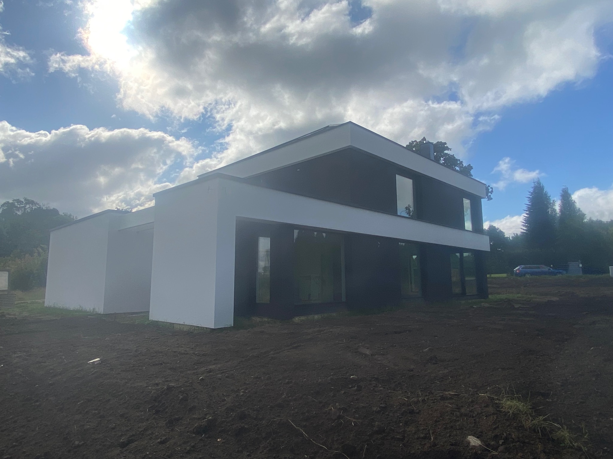 Nowoczesny dom z płaskim dachem, wykończony w bieli i czerni, na tle pochmurnego nieba i świeżo przekopanej ziemi.