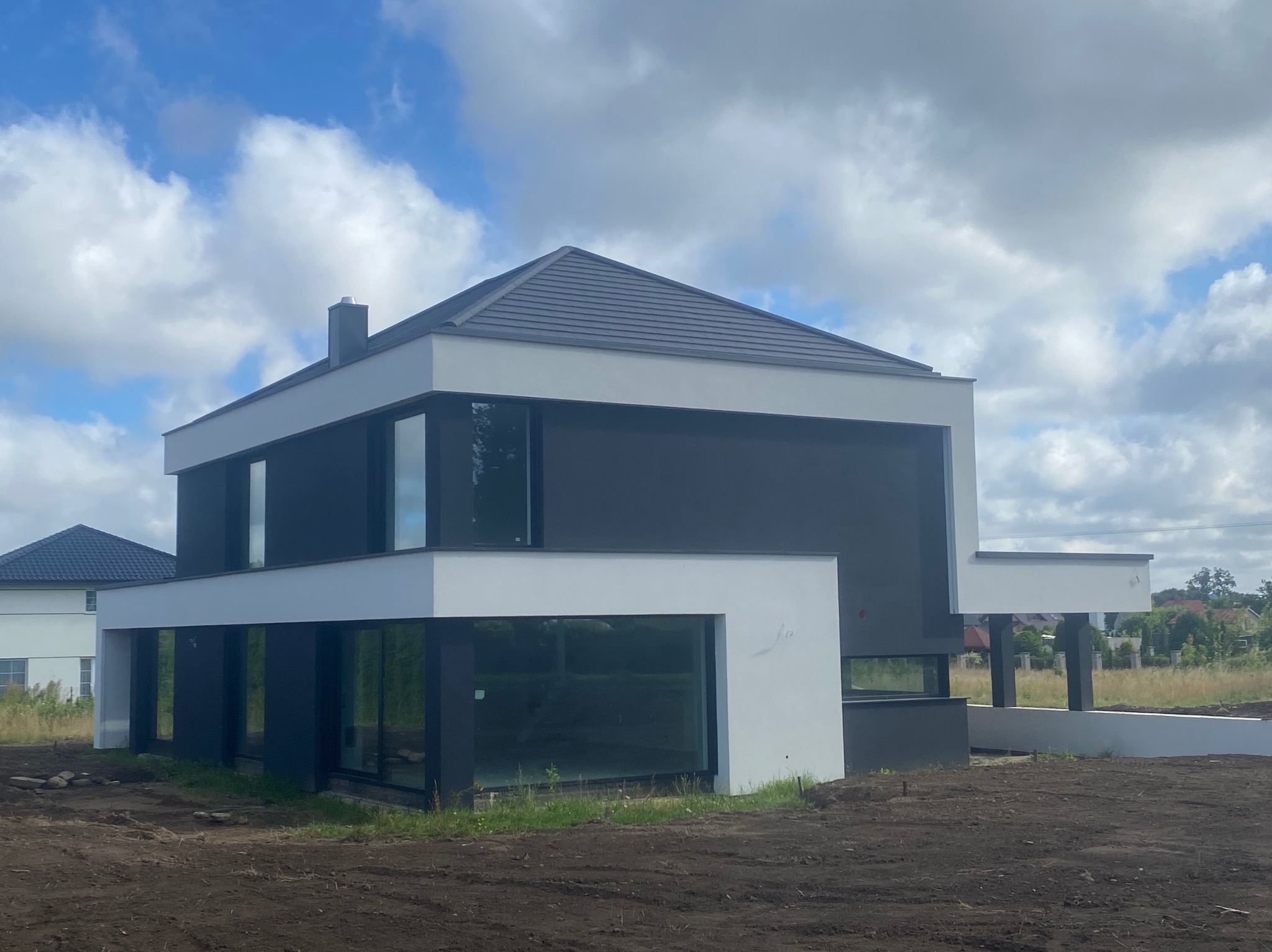 Nowoczesny, dwukondygnacyjny dom z płaskim dachem, wykończony w bieli i antracycie, z dużymi przeszkleniami, na działce z przygotowanym terenem pod ogród.